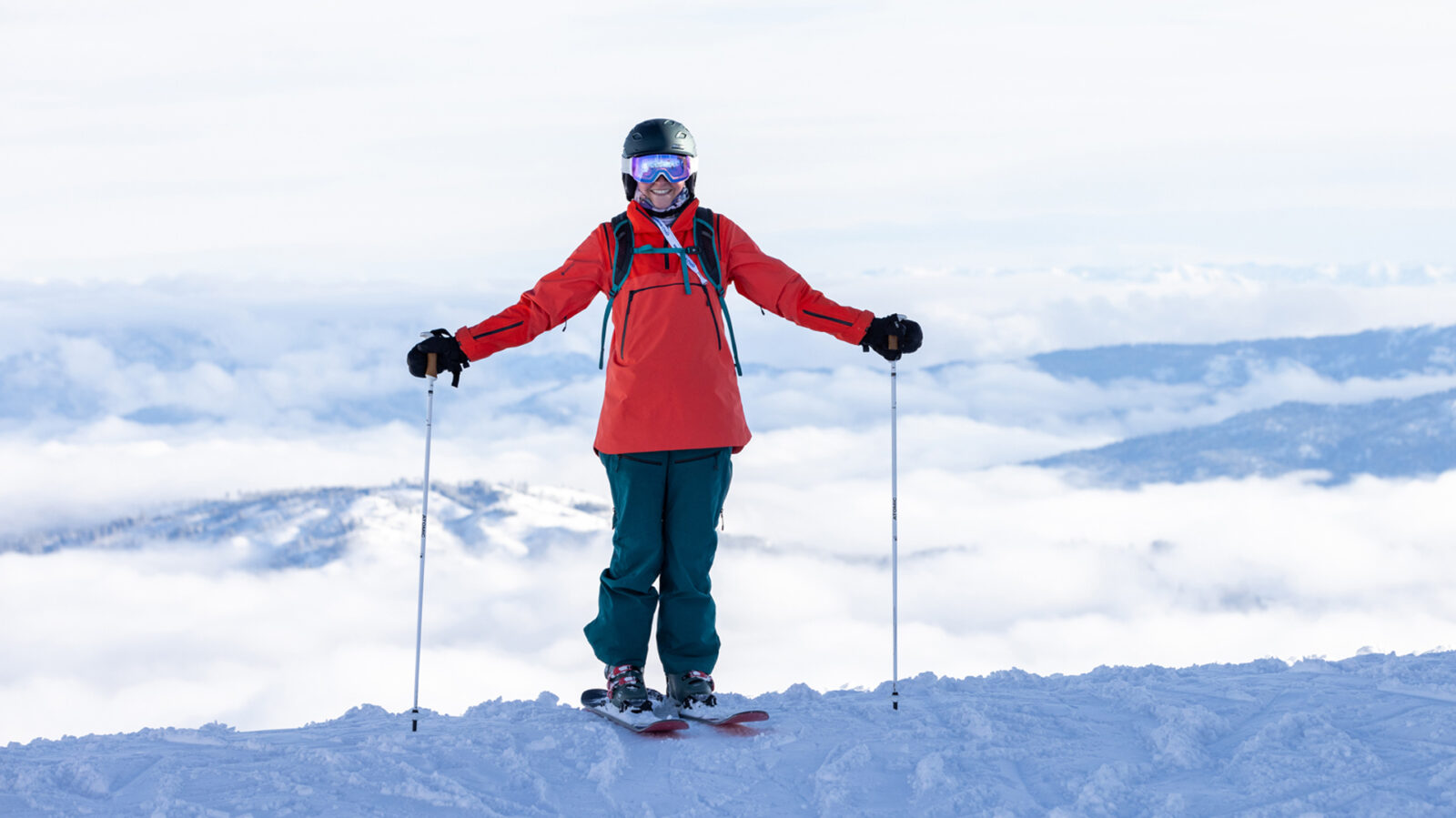 Smiling Adult Female Wearing Red Jacket Green Pants Backpack And Blue Lens Goggles Standing Arms Out With Inversion Background At Bogus Basin Mountain Recreation Area Winter2021 Photo By Luke Tokunaga 1920px1080px
