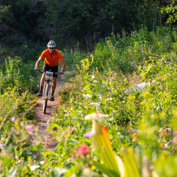 Male Mountain Biker Wearing Orange Jersey Rides On Deer Point Trail With Brush And Wildflowers In The Foreground Summer 2021 At Bogus Basin Mountain Recreation Area Photo By Luke Tokunaga 1920px1080px
