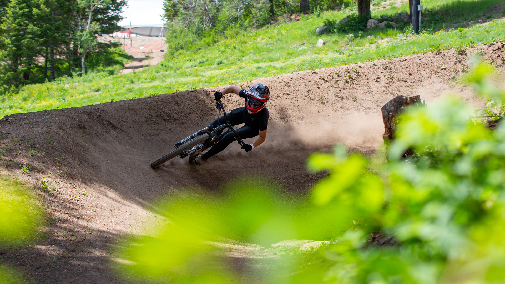 Mountain Biker Avery Riding A Berm On Fox Trot Basin Gravity Park Bike Trails With Green Bush In Foreground Summer 2021 At Bogus Basin Mountain Recreation Area 1920px1080px