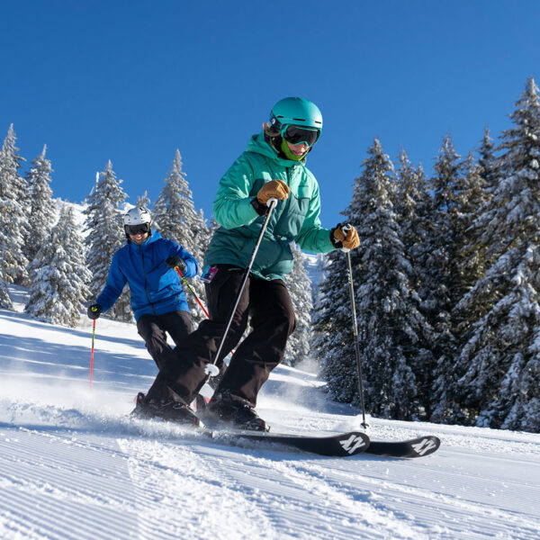 Adult Male And Female In Blue And Green Jackets Skiing Down Groomed Run On Bluebird Day Smiling Winter At Bogus Basin Mountain Recreation Area 1920px1080px