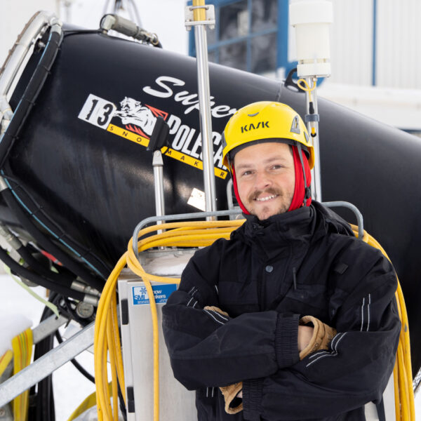 Bogus Basin Snow Making Male Employee With Yellow Helmet On Smiling In Front Of Black Pole Cat Snow Making Machine Next To Old Coach Chairlift Winter 2022 At Bogus Basin Mountain Recreation Area Photo By Luke Tokunaga 1920px1080px